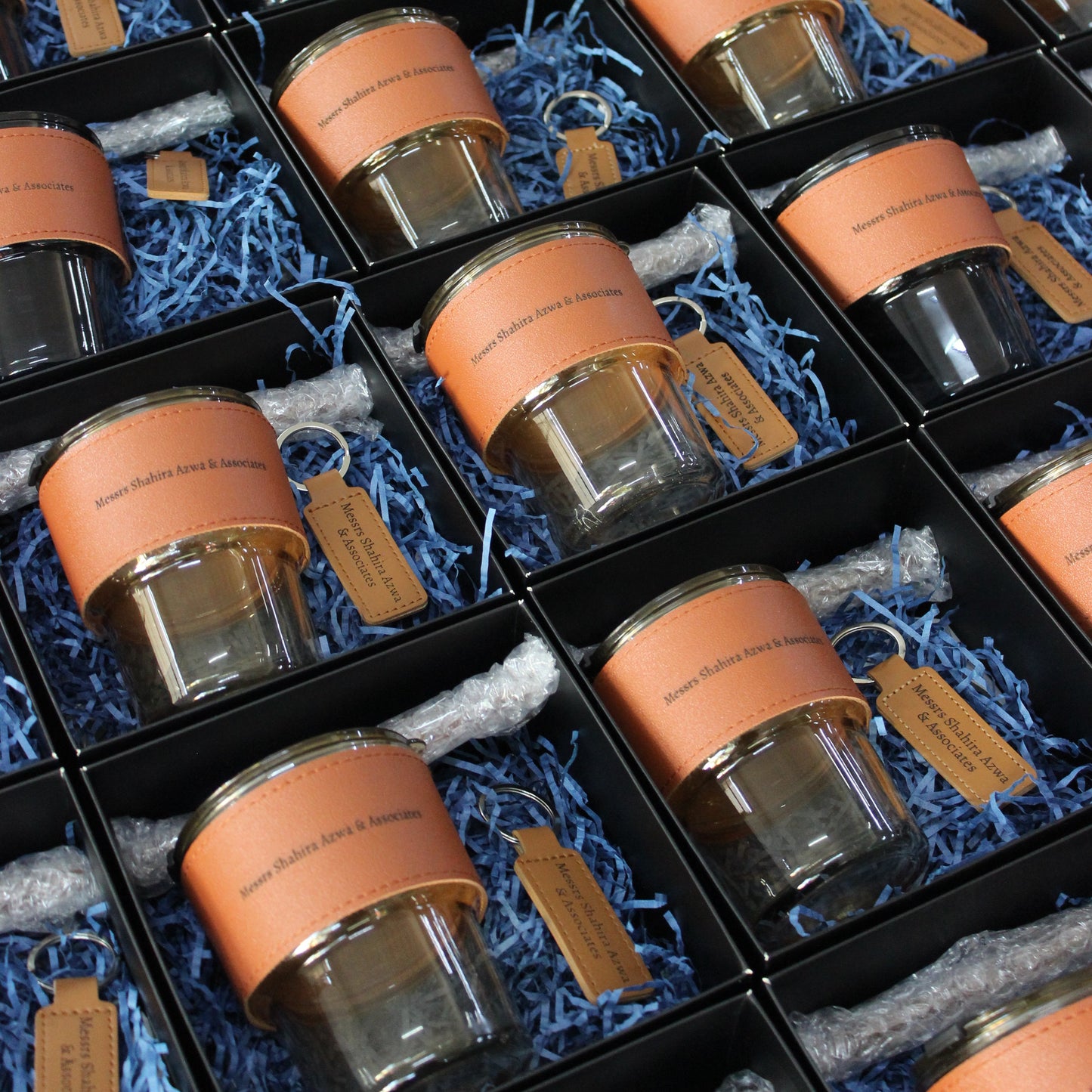 The image displays several corporate gift sets packaged in black boxes with shredded paper inside. Each set appears to contain a glass jar with a wooden stand and a keychain, though the keychains are not visible in the image.